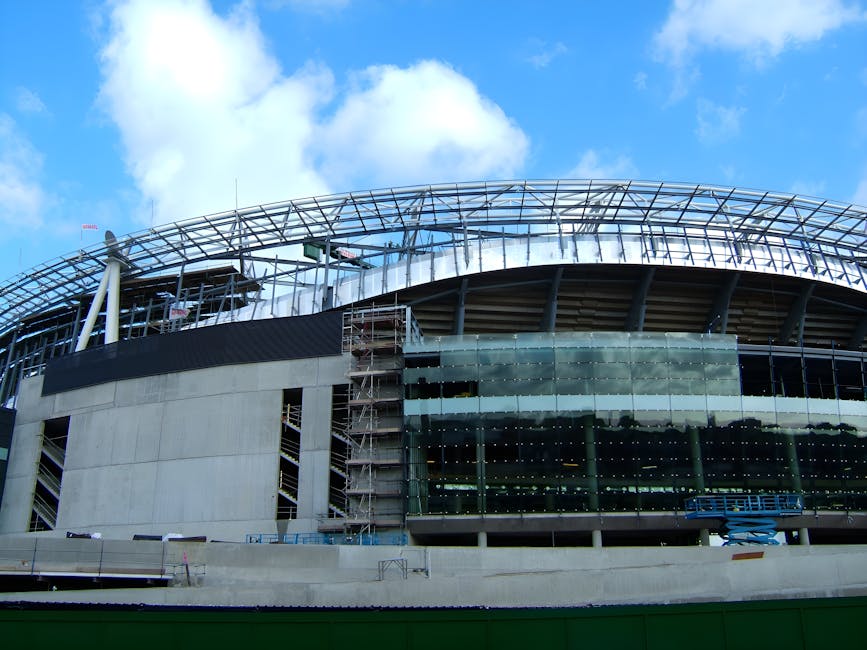 An empty football stadium with a well-maintained grass pitch, surrounded by red seating sections and modern white structural supports overhead. The seating area appears clean and orderly, with no visi