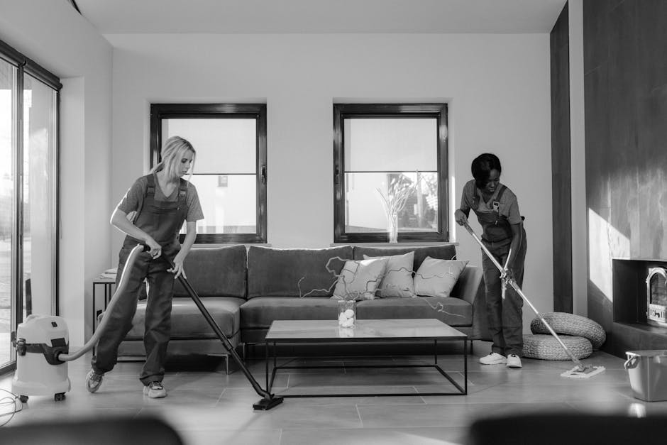 Two cleaning professionals from Cleaners Islington are performing surface cleaning and deep cleaning in a modern living room. One worker on the left uses a vacuum cleaner equipped with a hose attachment to clean the wooden floor, while the other on the right employs a mop to clean the tiled area near a grey wall. The room features a sofa with cushions, a glass-top coffee table with a small decorative item, and large windows allowing natural light to brighten the space. Cleaning supplies and equipment are visible, emphasizing a thorough approach to hygiene and maintenance aligned with domestic cleaning practices. The scene showcases meticulous surface cleaning on both wood and ceramic surfaces to ensure high standards of cleanliness and sanitisation.