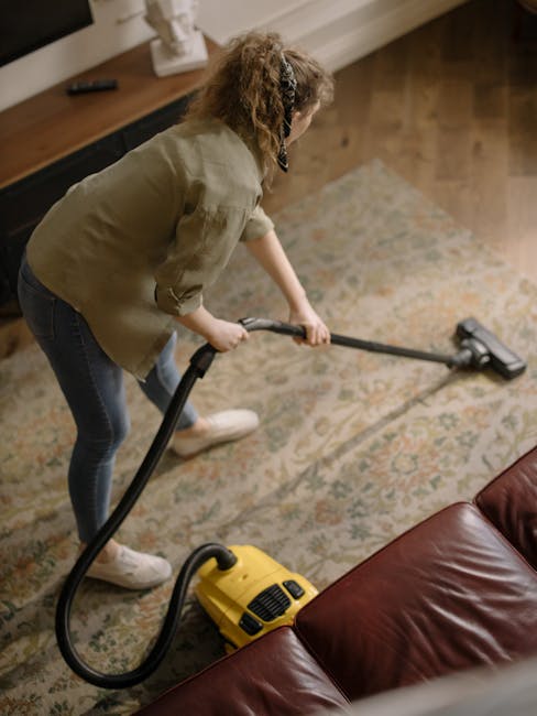 A woman wearing casual clothing and white sneakers is engaged in surface cleaning within a living room, using a yellow vacuum cleaner with a black hose attachment. She is performing deep cleaning on a patterned, multi-colored area rug situated on a wooden floor. The room features a brown leather sofa in the foreground and a wooden sideboard along the wall, which holds a white decorative item and possibly other small objects. The space is well-lit with natural light, highlighting the cleanliness and hygiene of the environment. This image exemplifies domestic cleaning services provided by Cleaners Islington for carpet and surface sanitisation, emphasizing meticulous maintenance of living spaces.