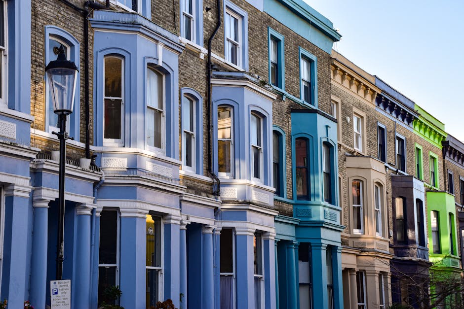 Image showing a row of Victorian-style terraced houses with decorative bay windows and painted facades in various colors, including light blue, teal, beige, black, and bright green, under natural daylight. The buildings feature brick and stucco exteriors with detailed cornices and window frames. A black streetlamp is visible in the foreground, standing on a sidewalk with a parking sign nearby. The scene depicts a clean, residential street in Islington, with well-maintained facades and no visible dirt or wear, representing a tidy urban environment. This setting exemplifies the type of properties that [COMPANY_NAME] provides professional cleaning services for, including end of tenancy cleaning and surface cleaning in the Upper Street Angel area of Islington.