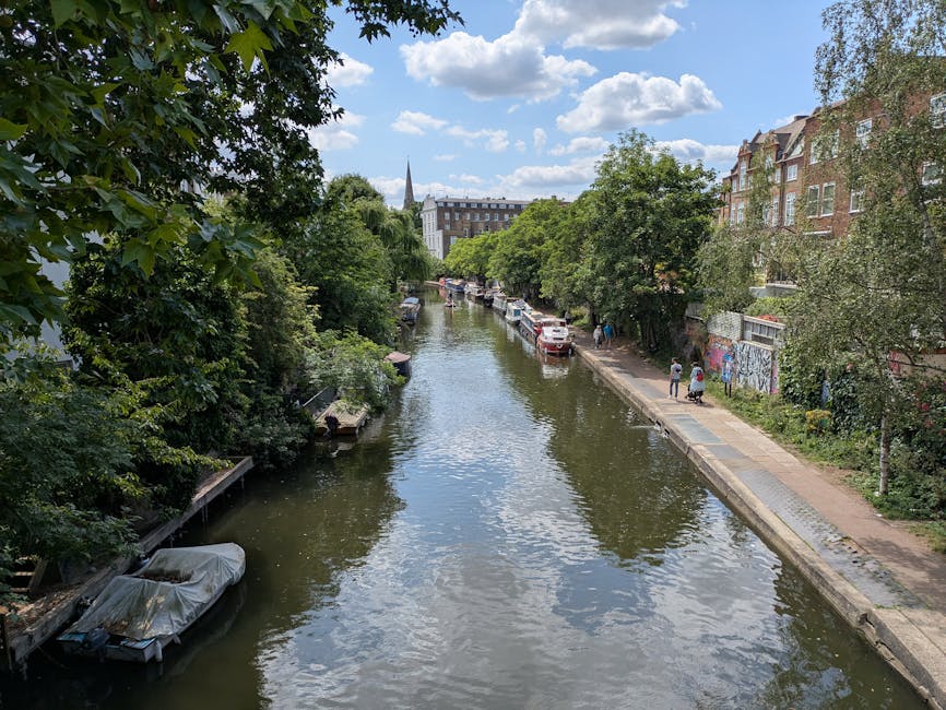 A scenic view of a narrow canal lined with lush green trees on both sides, reflecting the partly cloudy sky and nearby buildings. Several boats, some covered with protective tarps, are docked along the water's edge, while a paved footpath runs parallel to the canal with a few pedestrians walking. On the right, there are multistory brick buildings with large windows and front gardens, and a colorful mural is visible on a wall near the sidewalk. The scene is well-lit with natural daylight, highlighting the cleanliness and tranquility of the area, consistent with a well-maintained urban environment. This image exemplifies the peaceful, maintenance-free appearance typical of residential and commercial areas that benefit from professional cleaning services, as offered by Cleaners Islington, to preserve hygiene and aesthetic appeal.
