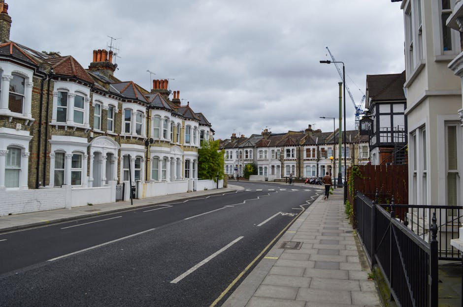 A quiet street scene in Barnsbury N1, Islington, featuring rows of Victorian terraced houses with white and brick facades, bay windows, and chimneys. The paved sidewalk runs alongside black metal railings and is bordered by residential buildings, some with small front gardens. The street is lined with parked cars, and a single person is walking along the sidewalk. Overhead, there are streetlights and construction cranes visible on the cloudy sky. The area appears well-maintained and clean, with no visible debris or dirt, reflecting a tidy urban environment consistent with professional cleaning standards. Cleaners Islington offers expert domestic cleaning and surface cleaning services to keep such residential areas hygienic and well-kept.