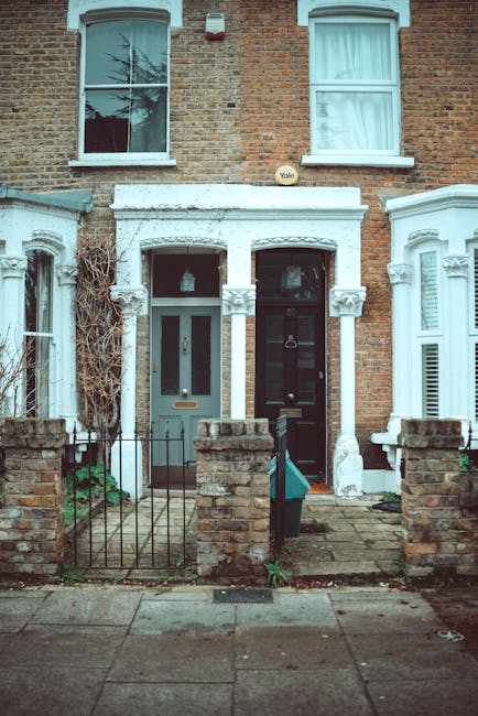 Photograph of the front doors of two residential terraced houses in Islington, featuring brick facades and decorative white window and door trims. The left house has a grey door with a glass panel and a brass mail slot, while the right has a black door with a brass knocker and letterbox. Both entrances have small paved front yards separated by low brick walls with metal railings, and the yard on the left includes a narrow gate. The upper floor windows are white-framed, with one displaying a partially drawn curtain and the other reflecting a bare tree. The overall appearance is clean and well-maintained, consistent with residential properties near Highbury Islington station. The image is associated with Cleaners Islington, showcasing the importance of surface cleaning and maintaining hygiene in residential settings.