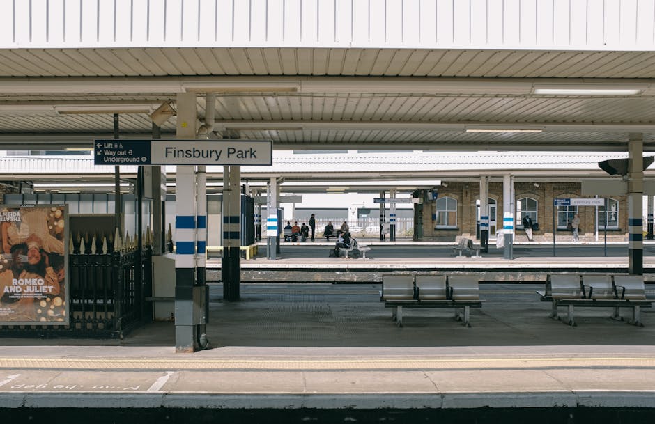 The Finsbury Park train station platform during daytime, featuring a clean and well-maintained concrete surface with visible tactile paving strips near the edge for safety. The platform has metal benches arranged for waiting passengers, and metal poles support a white, tiled canopy roof that provides shelter. In the background, there are a few commuters sitting or walking, with brick station buildings and signage visible. The lighting is even, highlighting the neatness of the area, which is indicative of thorough cleaning and maintenance by professional services such as Cleaners Islington, as referenced on the page for house cleaning near Highbury Islington station.