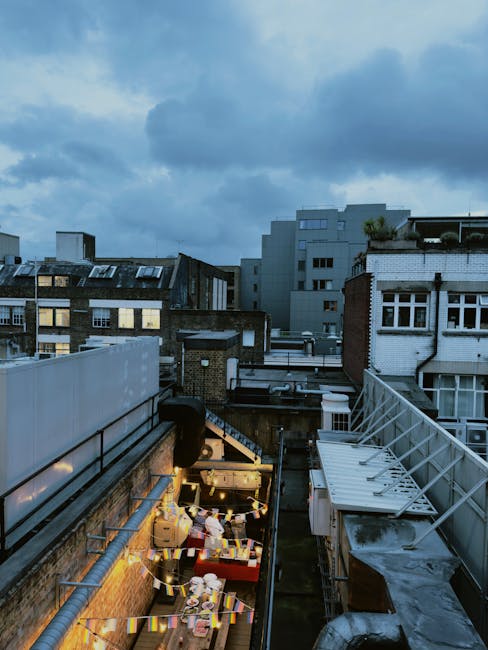 View of a cityscape from a rooftop during dusk, featuring a row of surrounding buildings with mixed materials including brick, glass, and metal. The sky is partly cloudy with a bluish hue. In the foreground, part of a terrace is visible with string lights and various items on a table, suggesting an outdoor area. Clean, tidy surfaces and well-maintained structures are apparent, with lighting highlighting the outdoor space. The image reflects an urban environment where residents might undertake or benefit from professional cleaning services, as offered by Cleaners Islington, especially for emergency or same-day domestic cleaning needs in Islington flats.