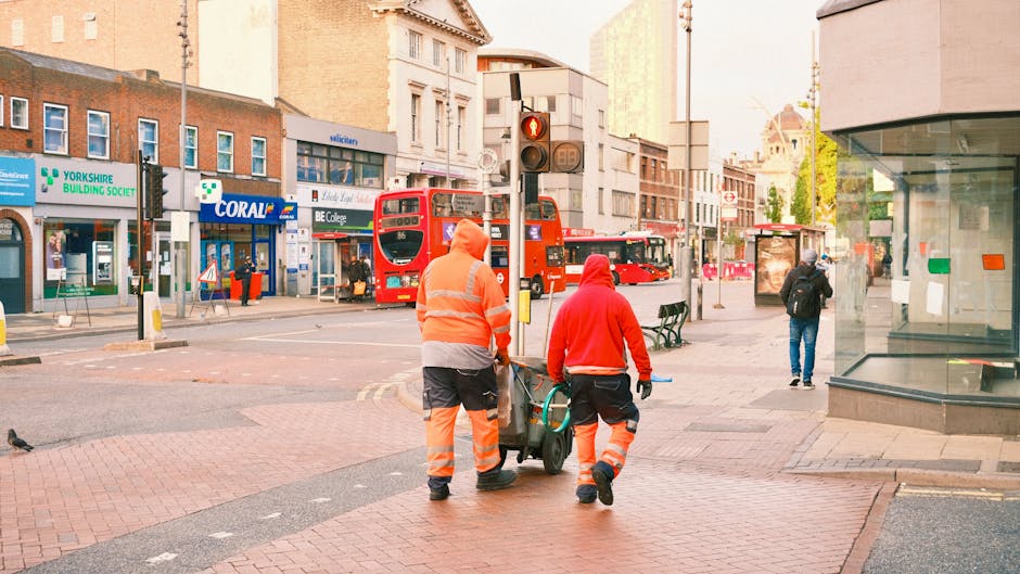 Two street cleaners wearing orange and red high-visibility uniforms are pushing cleaning equipment along a paved city sidewalk in Islington. The background features a busy urban scene with a red double-decker bus, storefronts including Yorkshire Building Society and Coral, and a bus stop. The area is well-lit with natural light, and the surfaces, including the pavement and shop windows, appear clean and well-maintained, reflecting routine surface cleaning and attention to hygiene, as part of the neighbourhood’s urban maintenance. This image illustrates community cleaning efforts aligned with professional hygiene standards described on the 'What to know about hidden cleaning charges in Islington' page by Cleaners Islington.